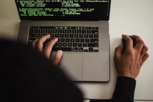 em primeiro plano, o capuz de um moletom desfocado. Depois, um computador da Apple sendo usado pela pessoa do capuz com a tela mostramdo códigos que representam ataques cibernéticos.