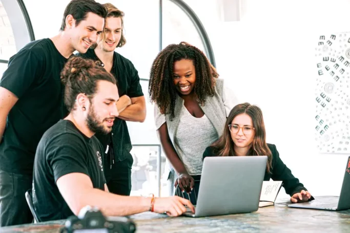 mercado-de-ti-pexels-canva-studio-3153201 Imagem de uma sala com fundo branco. Existe uma mesa com um notebook prateado e, ao redor do computador, cinco pessoas olhando para o conteúdo. São pessoas diversas, duas sentadas e três em pé, homens e mulheres que representam pessoas do mercado de ti.