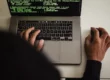 em primeiro plano, o capuz de um moletom desfocado. Depois, um computador da Apple sendo usado pela pessoa do capuz com a tela mostramdo códigos que representam ataques cibernéticos.
